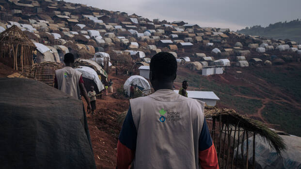 Zwei Mitarbeiter von Aktion gegen den Hunger von hinten in der Nähe eines Geflüchtetencamps in der Demokratischen Republik Kongo, auf den Westen das Logo