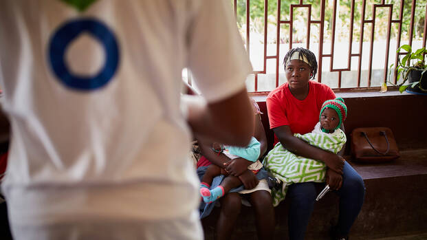 Eine Frau sitzt auf einer Bank mit einem Kind im Arm, während sie einer Erklärung eines unserer Mitarbeiterin in Haiti zuhört.