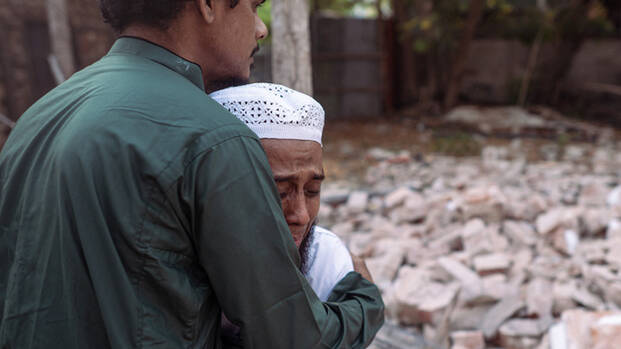 links: Zwei Männer umarmen sich fest nach dem Erdbeben in Myanmar. Im Hintergrund Steine eines eingestürzten Hauses. rechts: Menschen stehen an den Trümmern eines eingestürzten Gebäudes.