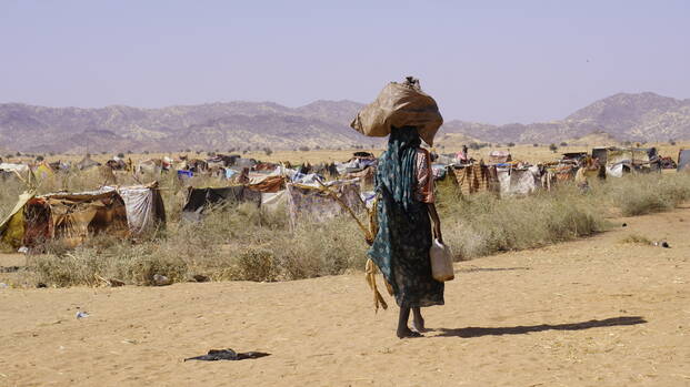 Eine Person im Sudan mit Gepäck auf dem Kopf geht durch eine trockene Landschaft, im Hintergrund ist ein provisorisches Lager zu sehen