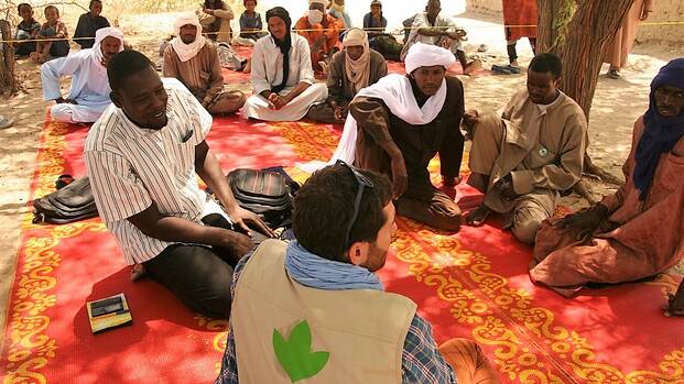 Helfer von Aktion gegen den Hunger sitzt mit einer Gruppe Männern und Kindern auf dem Boden.