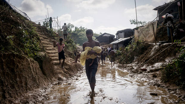 Junger Rohingya trägt in ein Tuch gewickeltes Baby durch knöcheltiefes Wasser in einem Flüchtlingscamp in Bangladesch