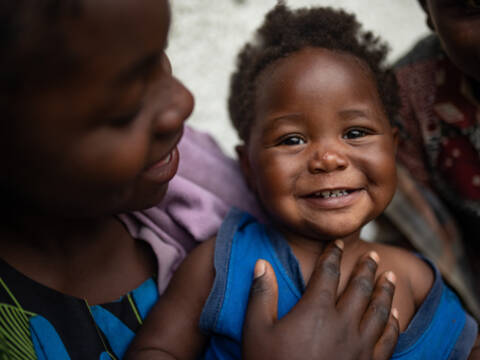 Ein kleines Mädchen aus der demokratischen Republik Kongo lacht in die Kamera, während es auf dem Schoß ihrer Schwester sitzt.